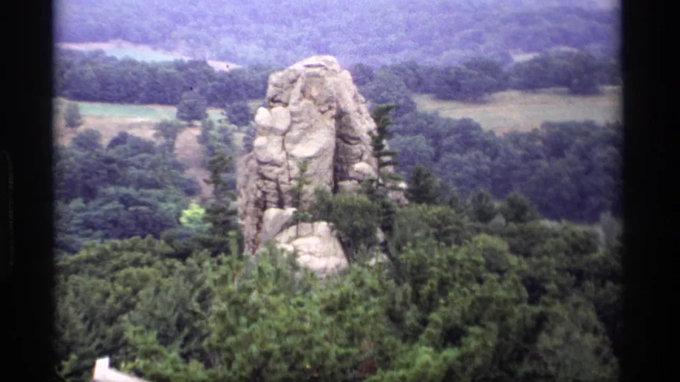 1969 Wisconsin home-movie frame showing a rock spire rising from thick foliage with wooded slopes behind it.