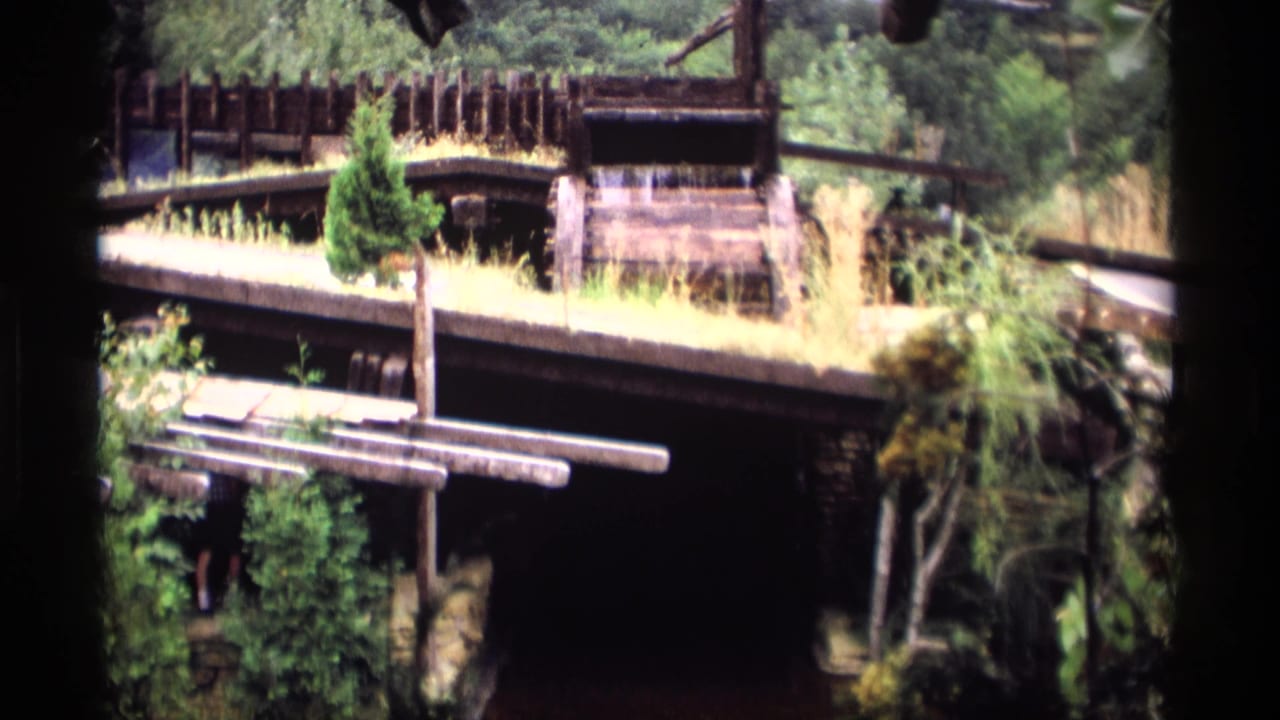 1969 Wisconsin 8mm frame of a rustic waterwheel setting surrounded by wood railings, trees, and overgrown plants.