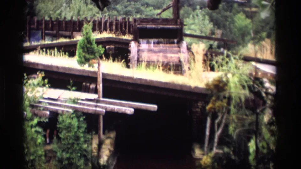 1969 Wisconsin 8mm frame of a rustic waterwheel setting surrounded by wood railings, trees, and overgrown plants.