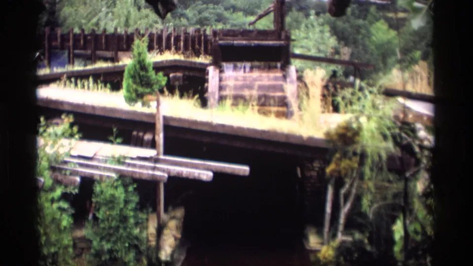 1969 Wisconsin home-movie frame showing a closer view of the waterwheel structure, railings, plants, and shaded water.