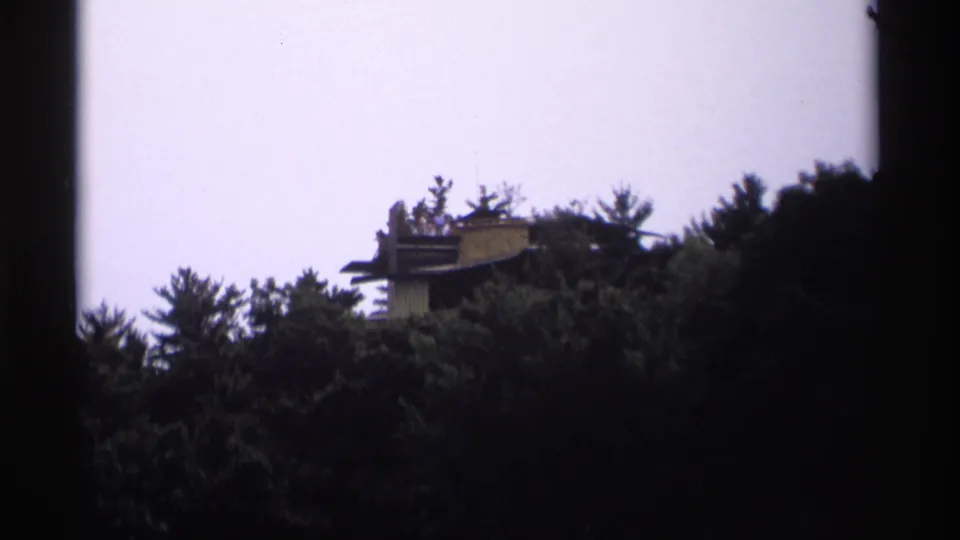 1969 Wisconsin 8mm frame showing a distant structure above dense evergreens, partly obscured by foliage.