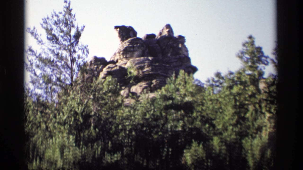 Layered rock formation rises above green trees against a pale sky.