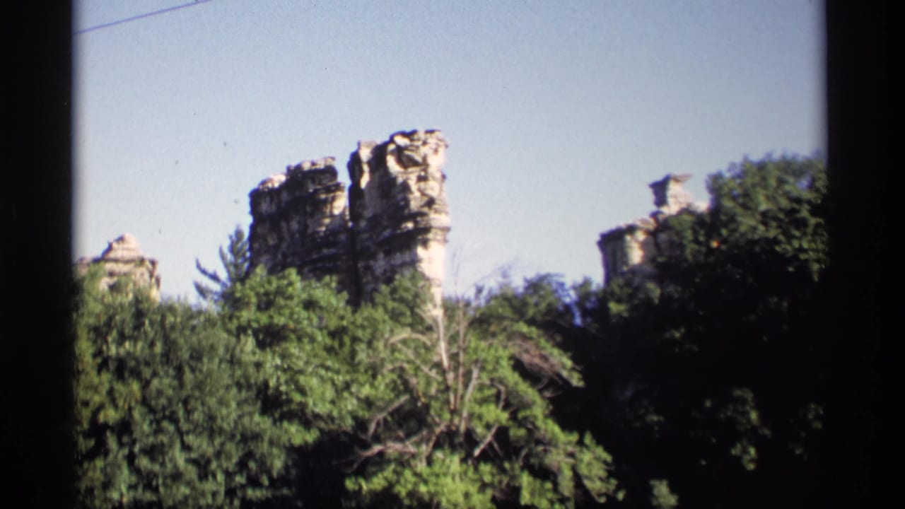 Layered rock formations rise above dense green trees under a pale blue sky.