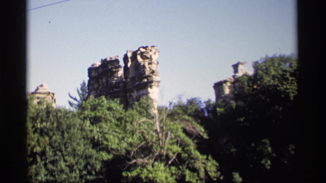 1969 Wisconsin 8mm view of tall, weathered rock outcrops partly obscured by dense green foliage.