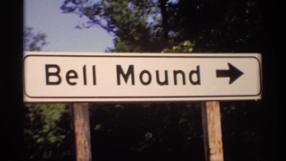 1969 Wisconsin frame showing a white Bell Mound directional sign with a right-pointing arrow, set against trees and sky.