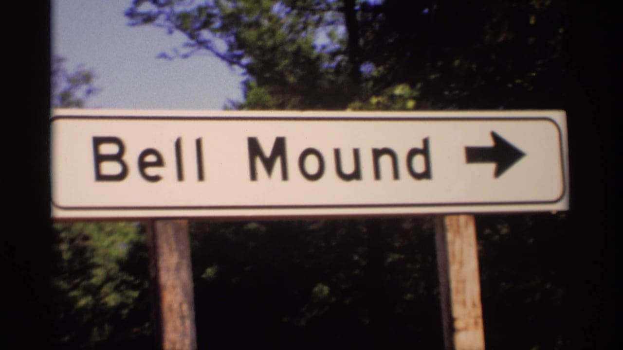 1969 Wisconsin 8mm frame showing a white Bell Mound directional sign with a black arrow, mounted on wooden posts near trees.