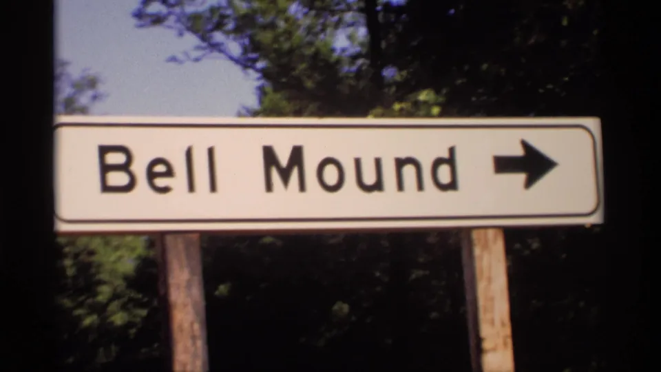 1969 Wisconsin 8mm frame showing a white Bell Mound directional sign with a black arrow, mounted on wooden posts near trees.