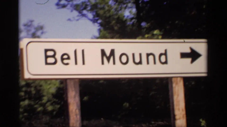 The Bell Mound sign remains centered in a 1969 Wisconsin home-movie frame, with a black arrow pointing right and trees behind it.