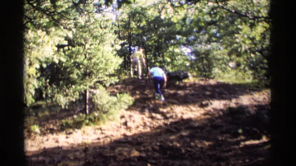 A 1969 Wisconsin home-movie frame shows two distant hikers moving through trees on uneven, sunlit ground.