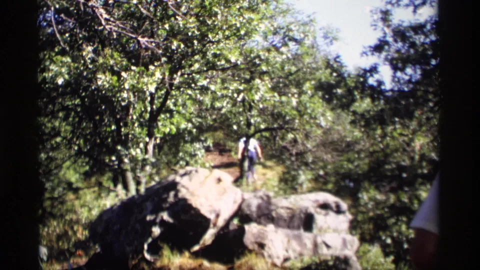 A 1969 Wisconsin home-movie frame shows a hiker near boulders on a wooded path, with bright leaves and brush around the trail.