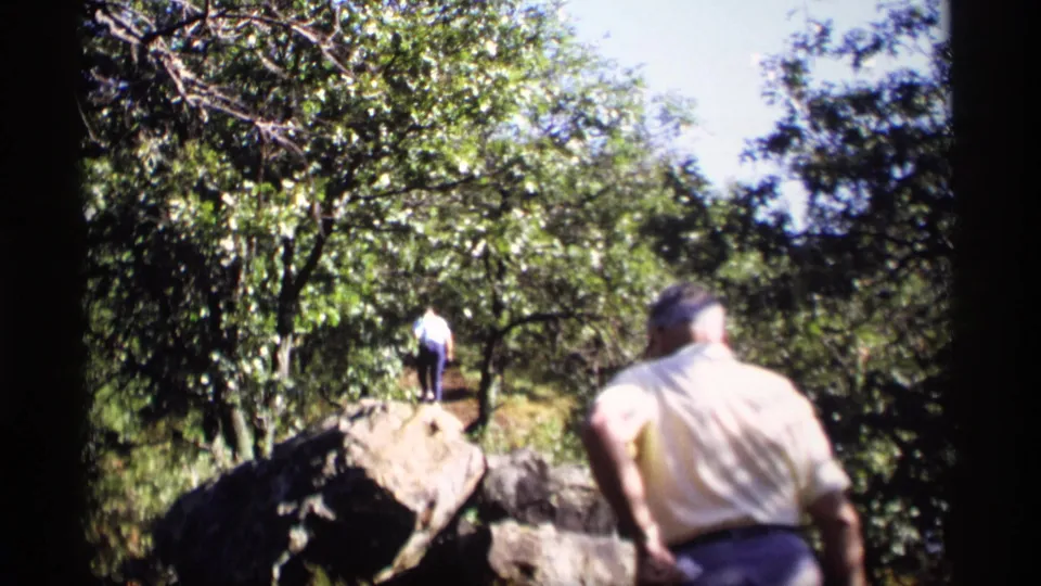 Two people appear in a 1969 Wisconsin hiking scene, with one close to camera and another farther ahead near large rocks and trees.