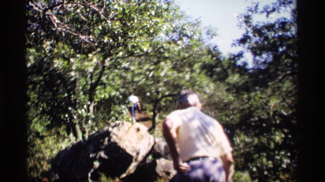 1969 Wisconsin 8mm frame showing an older man from behind on a rocky hillside, with another person farther up among green trees.