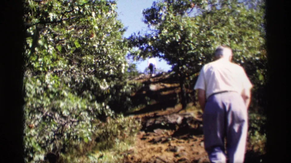The older man continues upward through a wooded Wisconsin hillside, seen from behind in soft 8mm color and dappled sunlight.