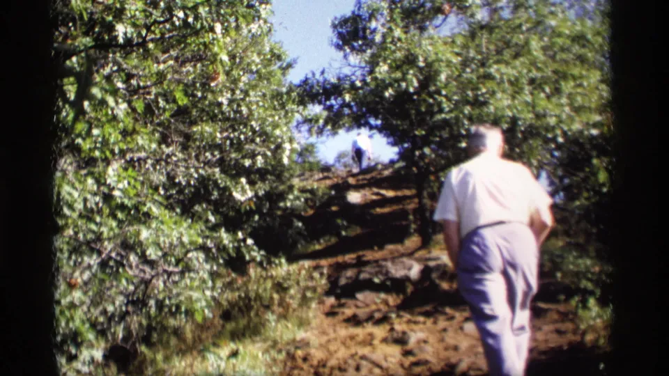 A handheld 8mm view follows the man from behind as he climbs a rocky, shaded hillside in Wisconsin, with another figure ahead.