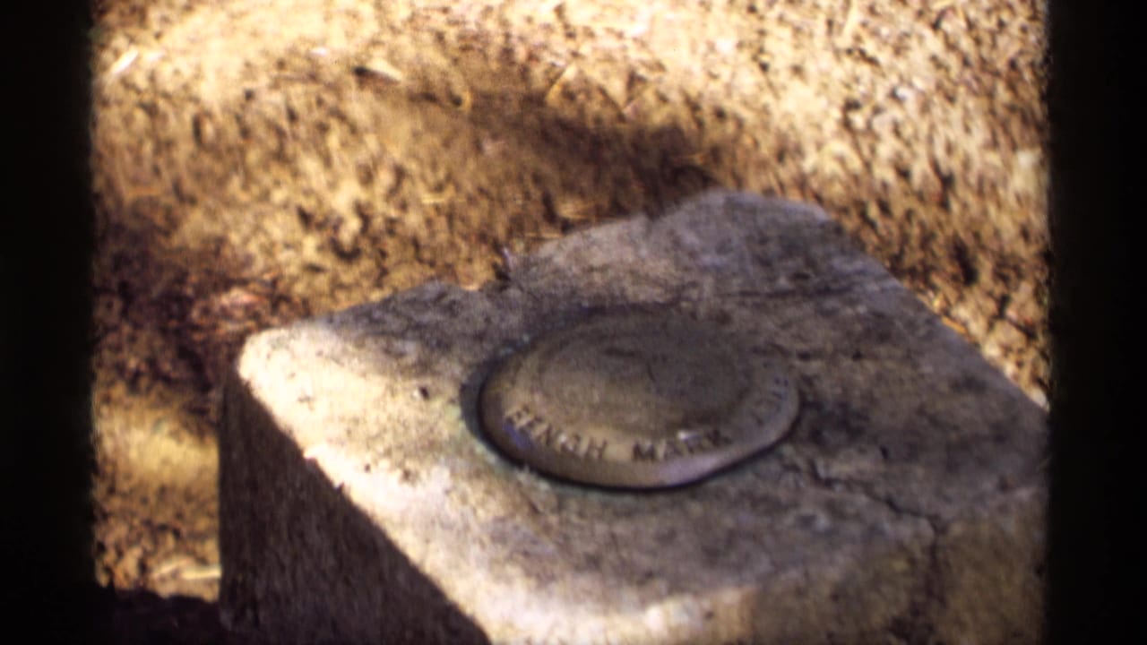 Bench mark plaque on a stone block with soil and shadow behind it.