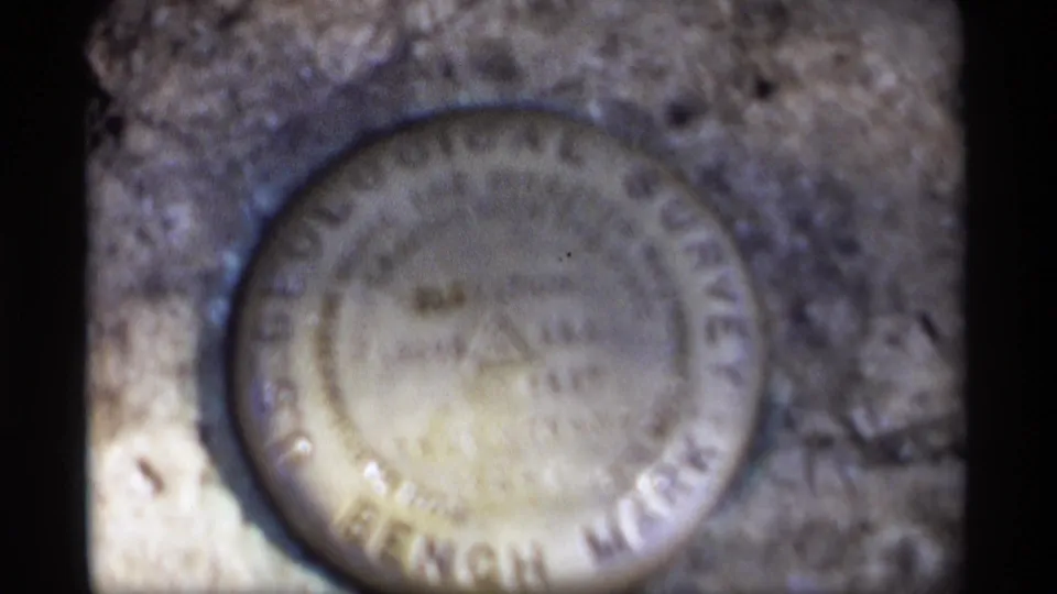 Final close 8mm frame of the U.S. Geological Survey bench mark, softened by focus and film grain on the stone pedestal.