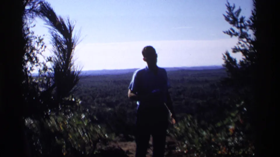 A silhouetted man in 1969 Wisconsin home-movie footage stands near brush and trees, looking out over a broad forested valley.