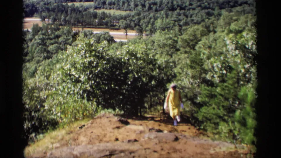 In rural Wisconsin home-movie footage from 1969, a woman in a yellow dress walks along a wooded dirt path below the hillside view.