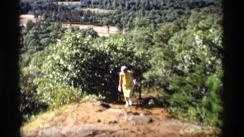 In 1969 Wisconsin home-movie footage, a woman in a yellow dress and white gloves walks up a dirt path bordered by trees.