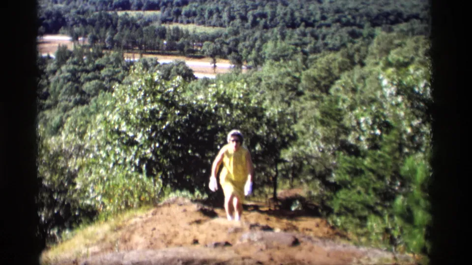 The woman is nearer to the camera on the rough hillside path, framed by dense greenery and distant landscape below.