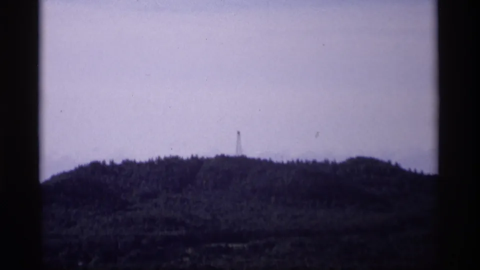 1969 Wisconsin home-movie frame showing a distant wooded ridge, pale sky, and a slender tower near the horizon.