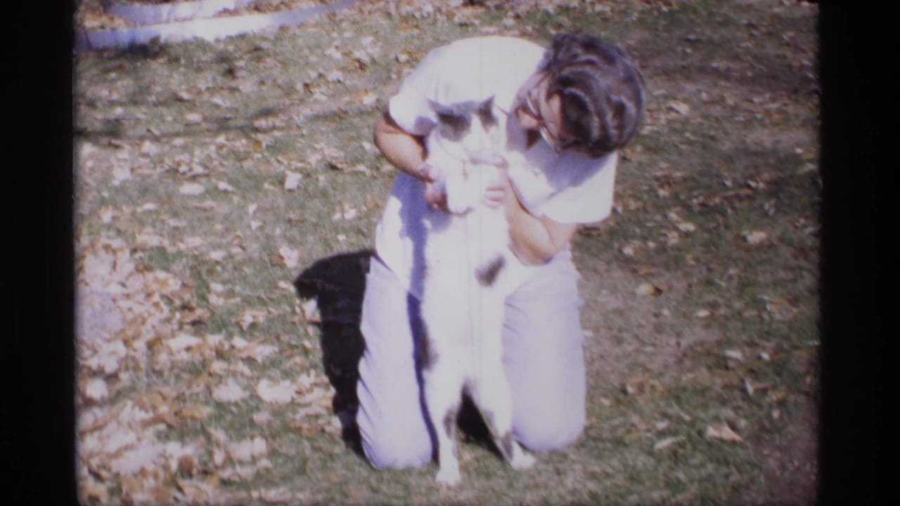 Woman kneeling on a lawn while holding a black-and-white cat upright.