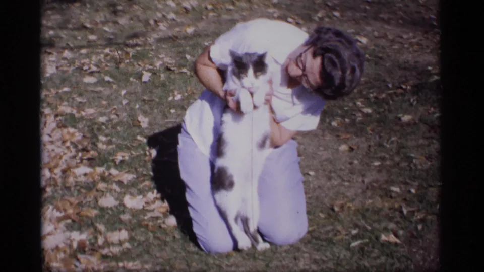 A kneeling woman holds the black-and-white cat close on a grassy Wisconsin lawn, with fallen leaves visible around them.