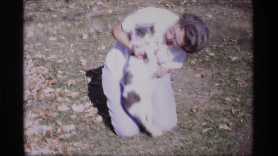 The woman crouches in bright sun as the black-and-white cat shifts in her hands on the autumn lawn.