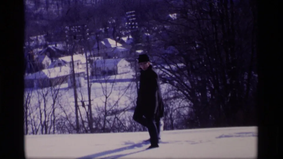 In Wisconsin 1969 footage, a man in a dark coat and hat stands on a snowy slope overlooking rooftops, trees, and distant buildings.