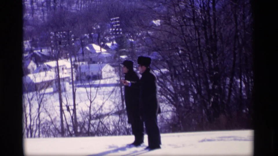 Two men in dark winter coats and hats stand on a snowy Wisconsin overlook, facing a townscape with bare trees and rooftops below.