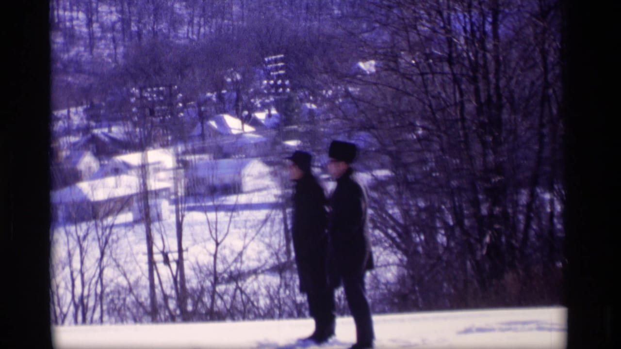 Two men in dark winter coats stand on a snowy hill above distant houses.