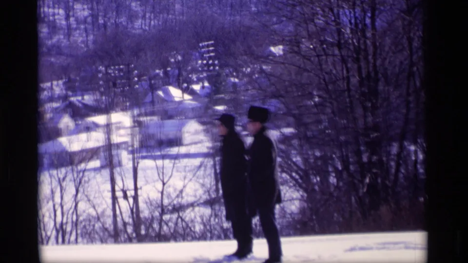 In Wisconsin 1969 home-movie footage, two men in dark coats and hats stand on snow with a residential neighborhood below.