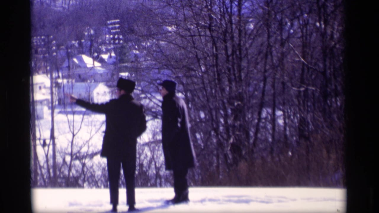 Two men stand in snow while one gestures toward the neighborhood below.