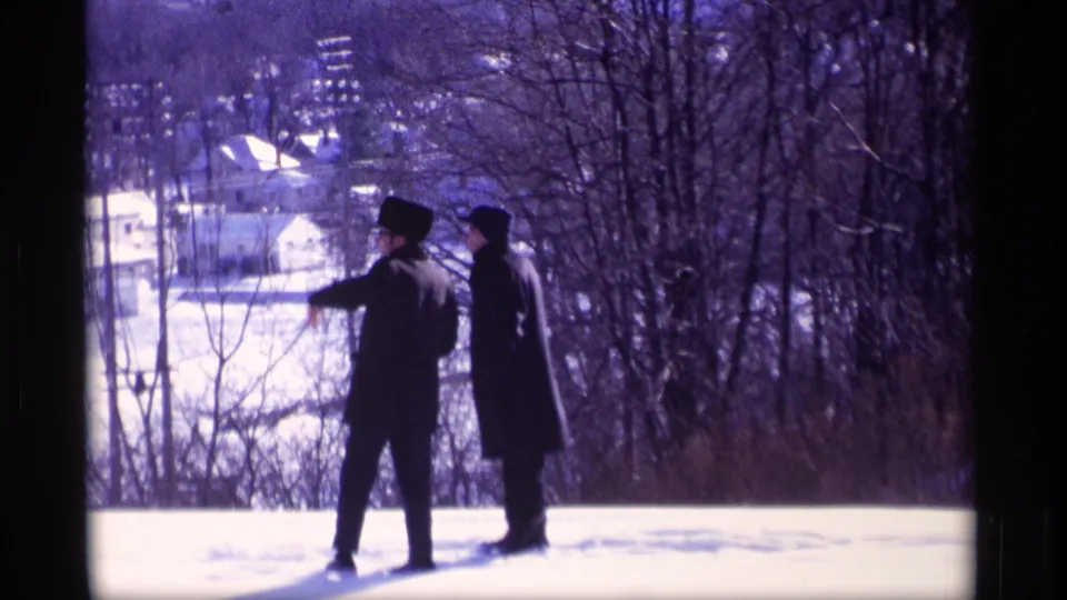 In a 1969 Wisconsin winter scene, two men stand on a snow-covered overlook framed by bare trees and distant homes.