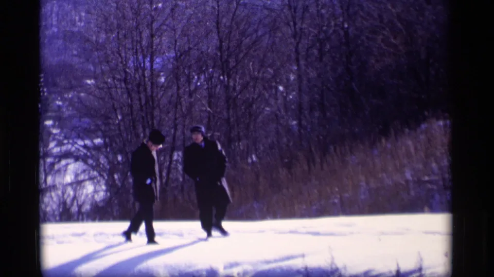 Two men in dark winter clothing walk together across a snowy Wisconsin hillside in vintage 8mm footage.