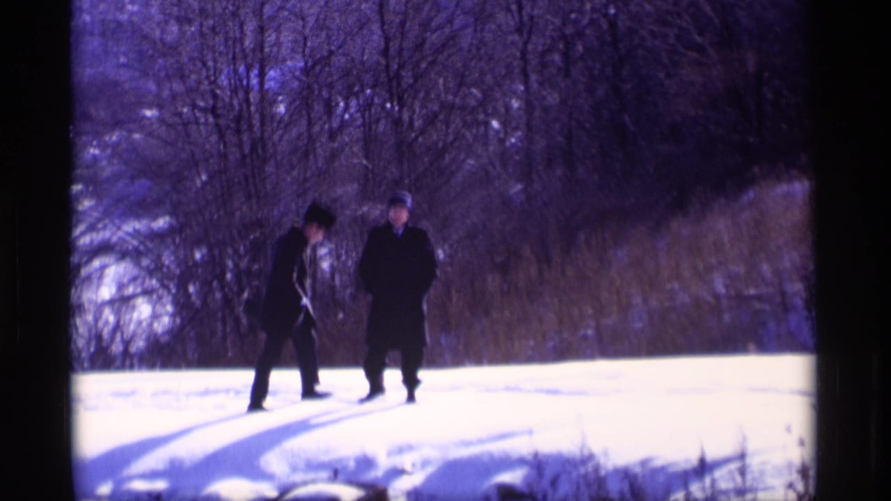 Two men in dark coats and hats walk on snow near bare winter woods.
