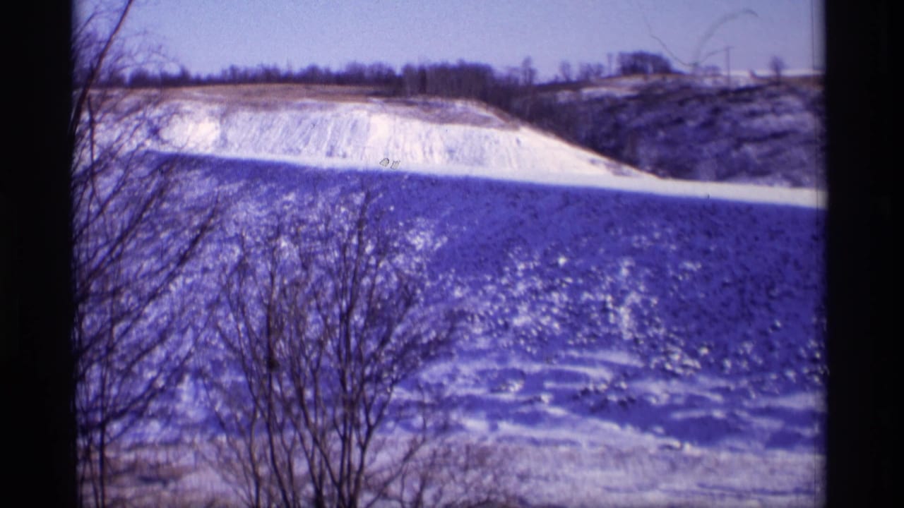 Snow-covered field and hillside with bare branches in the foreground.