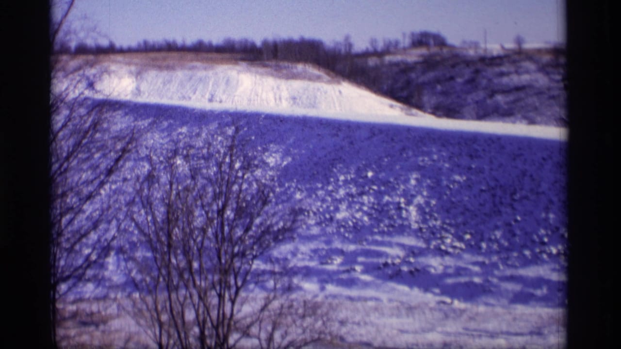 Vintage 8mm winter view in Wisconsin, with bare foreground trees crossing a sunlit snowy field and distant ridge.