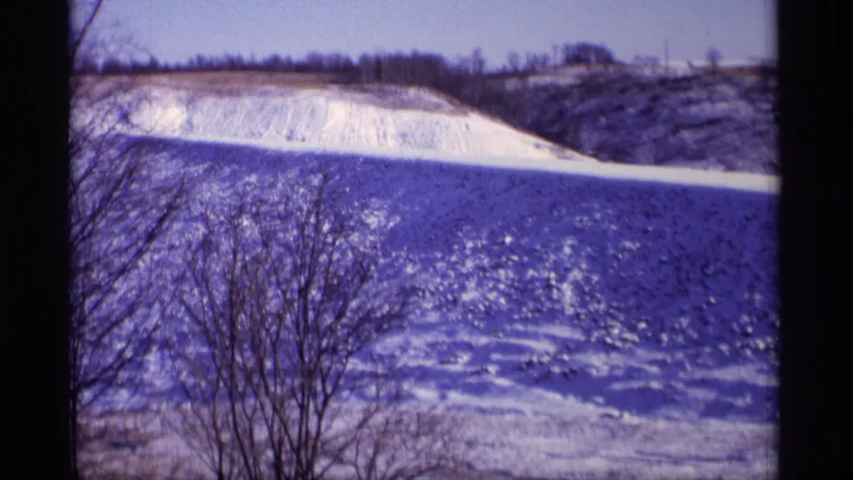 Vintage 8mm winter view in Wisconsin, with bare foreground trees crossing a sunlit snowy field and distant ridge.