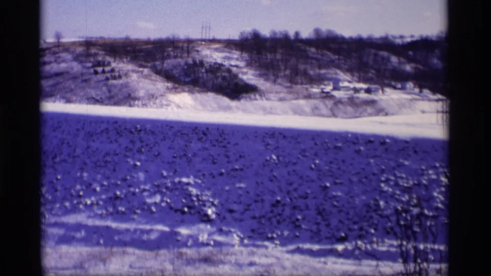 1969 Wisconsin home-movie frame showing an open snow-covered field, distant trees, small structures, and utility poles.