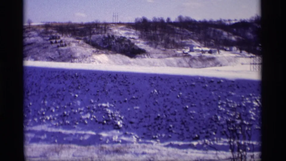 Vintage 8mm frame from Wisconsin showing a snowy field, bare treeline, utility poles, and small structures on the horizon.