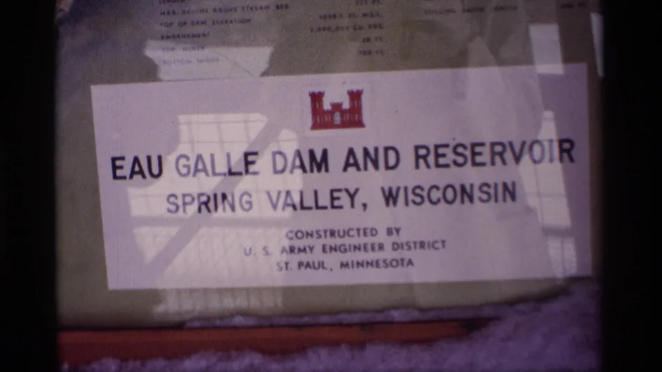 1969 Wisconsin 8mm frame showing the Eau Galle Dam and Reservoir sign for Spring Valley, with reflections across the glass.