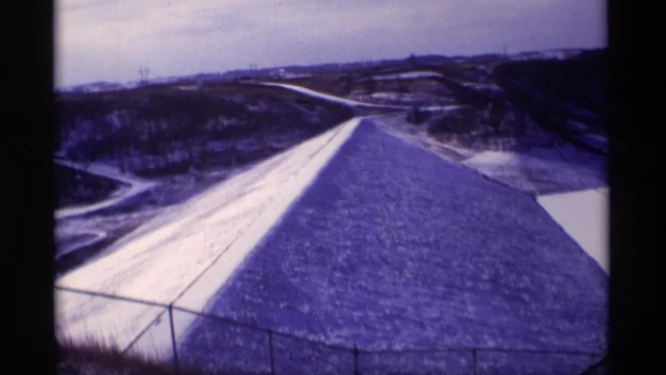 1969 Wisconsin 8mm frame showing the dam face from above, with a fence in the foreground and rural terrain in the distance.