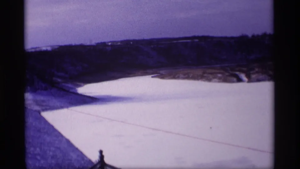 1969 Wisconsin 8mm frame showing a snowy embankment, frozen reservoir, dark shoreline, and low hills under a pale sky.