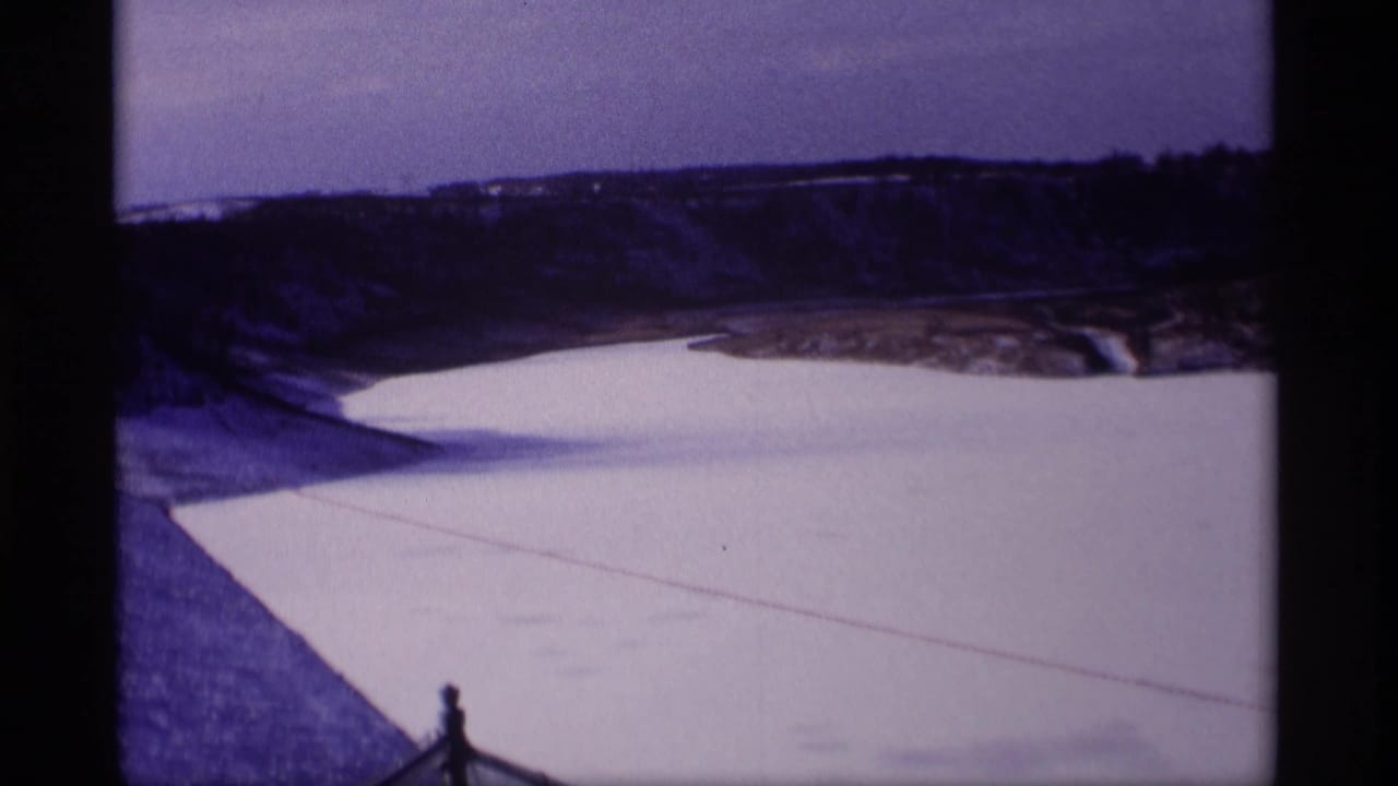 1969 Wisconsin home-movie view of a broad frozen reservoir bordered by a sloped snowy bank and distant wooded hills.