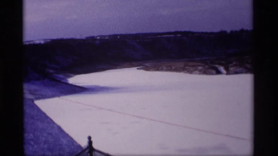 1969 Wisconsin home-movie view of a broad frozen reservoir bordered by a sloped snowy bank and distant wooded hills.