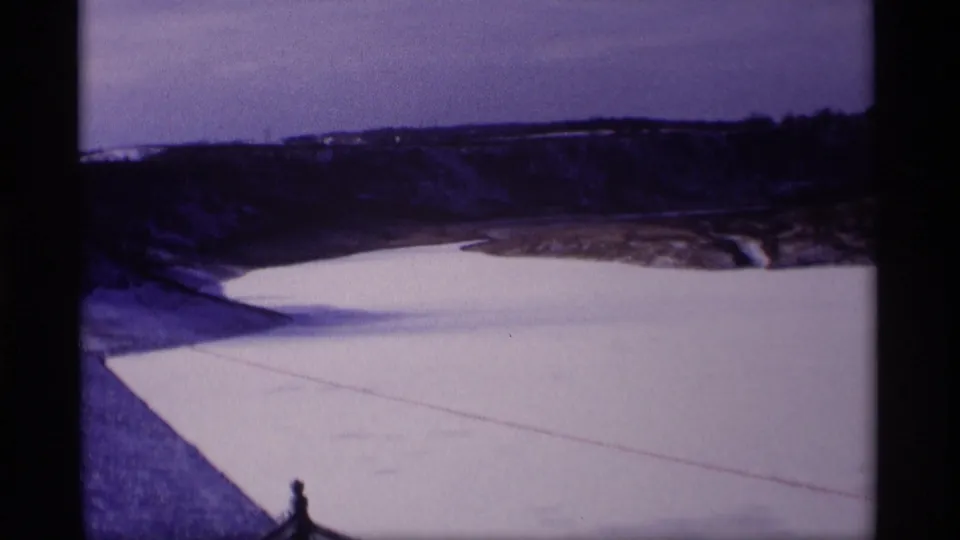 1969 Wisconsin 8mm still with a smooth frozen reservoir, snowy foreground slope, distant ridge, and pale winter sky.