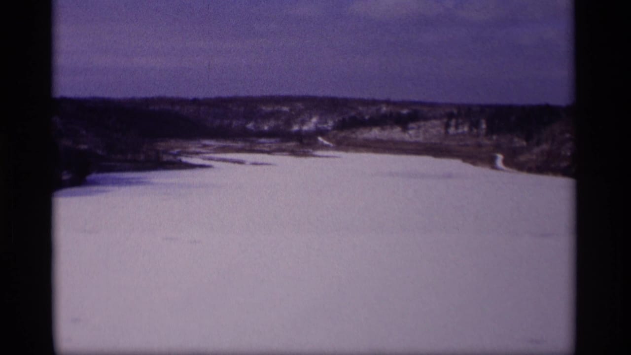 1969 Wisconsin 8mm frame showing a frozen lake or river valley with snow-covered banks, bare trees, and low hills beneath a muted sky.