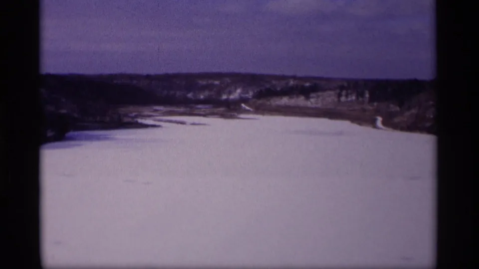 1969 Wisconsin 8mm frame showing a frozen lake or river valley with snow-covered banks, bare trees, and low hills beneath a muted sky.