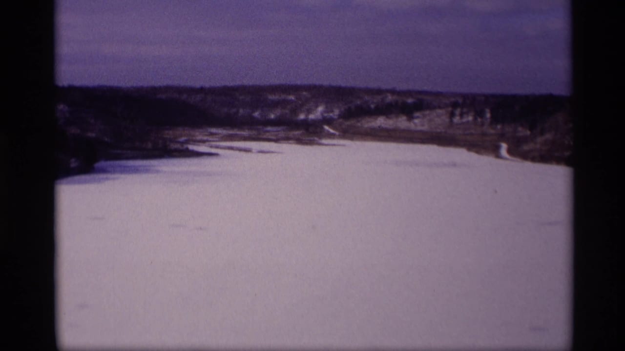 Wide frozen water surface with snow along the banks and low hills in the distance.